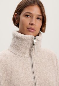 Fleece zip-jakke i lys beige med højt krave, der har en tekstureret, fluffy overflade og en sølv lynlås med en læder trækstang.