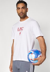 Λευκή βαμβακερή μπλούζα T-shirt με κόκκινο λογότυπο "LFC" και κείμενο, συνδυασμένη με γκρι παντελόνι. Το άτομο κρατάει μια πολυχρωμη μπάλα ποδοσφαίρου με σχέδια.