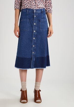 Kvinde iført en mid-lang blå denimnederdel med knapper foran og store lommer, kombineret med beige sokker og brune åbne ankelsko.