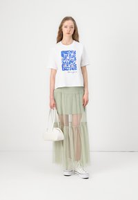 T-shirt graphique blanc avec un design floral bleu, accompagné d'une jupe superposée légère verte. La mannequin tient un sac à main blanc et porte des baskets blanches.