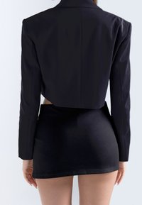Kvinna iklädd en svart kort blazer och en åtsittande svart minikjol, stående med ryggen mot kameran mot en ensam bakgrund.
