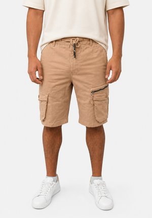 Uomo con pantaloncini cargo beige con tasche con zip e bottoni, scarpe da ginnastica bianche, in piedi su uno sfondo semplice.