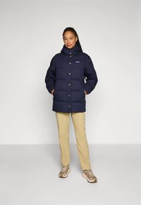 Marinblå vadderad jacka med hög krage, tryckknappar och framfickor. Kombinerad med beige byxor och ljusbruna skor. Slät tygstruktur.