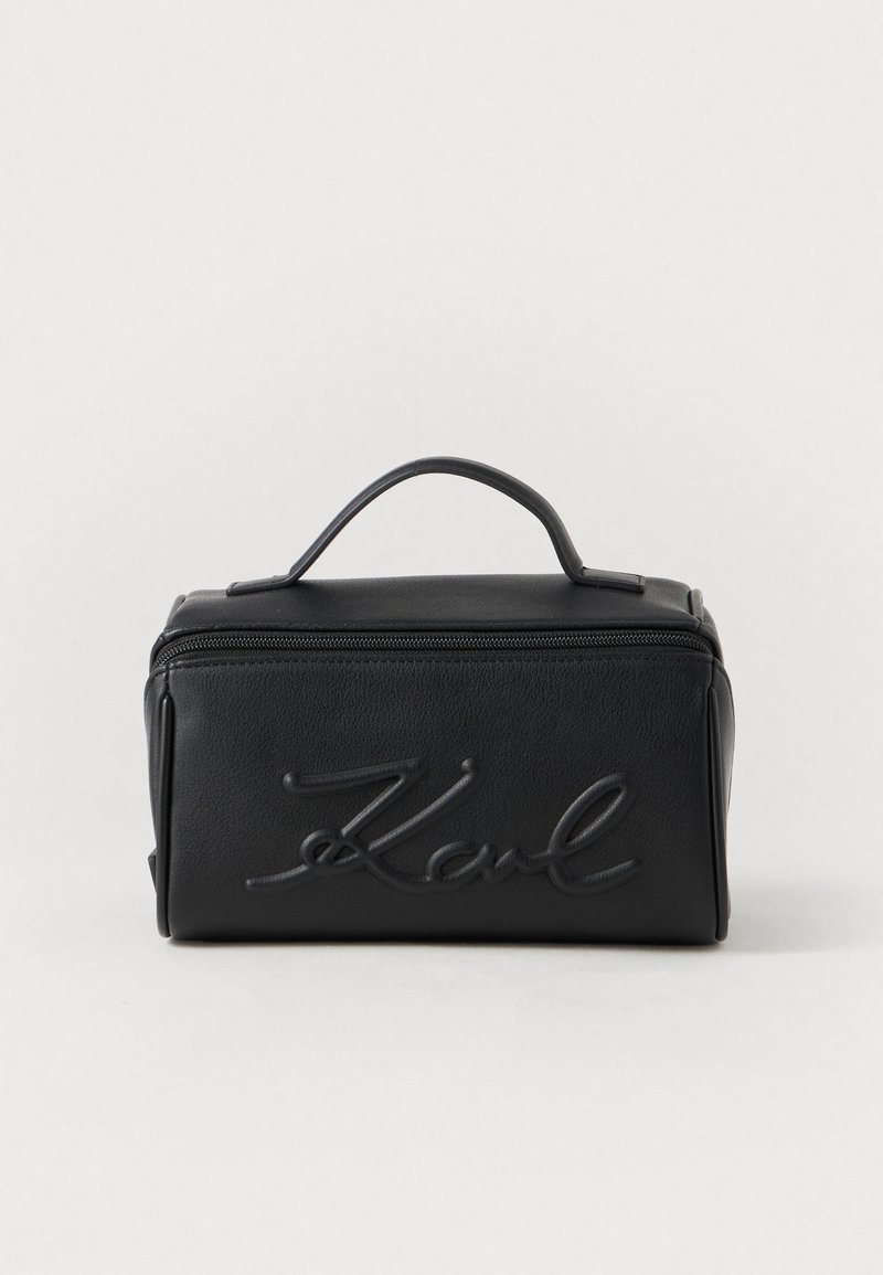 Estuche cosmético de cuero negro con un asa en la parte superior, logotipo grabado en la parte frontal, forma rectangular y cierre de cremallera. Textura suave, diseño minimalista.