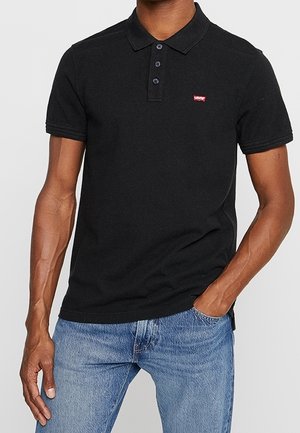 Homme portant un polo noir à manches courtes avec trois boutons et un petit logo rouge Levi's, associé à un jean bleu, main dans la poche.