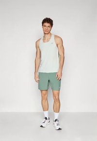 Ung mann iført en lys grå ermeløs treningstrøye, grønne shorts, hvite sokker og hvite joggesko, som står mot en ensfarget bakgrunn.