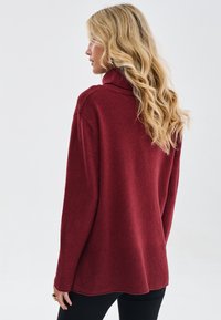 Bordowy sweter z golfem o luźnym kroju, z prążkowanym dołem i teksturowaną dzianiną. Długie rękawy i zaokrąglony tył dopełniają całość.