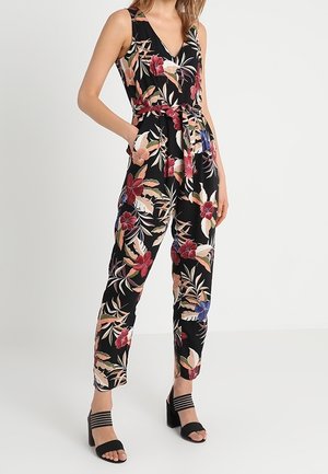 Femme portant une combinaison noire sans manches avec imprimé floral rouge, rose et bleu, taille cintrée et sandales à talons noires.