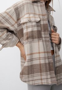 Brązowa i beżowa koszula oversize z tkaniny w kratę, wykonana z miękkiego materiału, posiadająca dwie kieszenie na piersi, zapięcie na guziki i luźny krój.