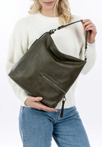 Green leather shoulder bag with a slouchy design, featuring a front zipper pocket and a detachable strap, held by a person wearing a white sweater.