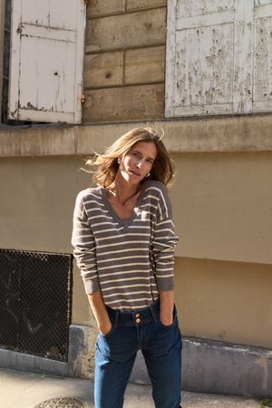 Femme aux cheveux mi-longs portant un pull rayé gris et blanc et un jean bleu, debout les mains dans les poches devant un mur usé.