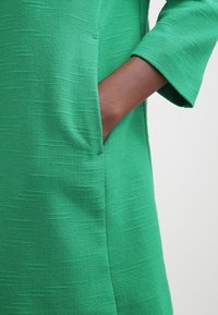 Zelené texturované šaty s přední kapsou a tříčtvrtečními rukávy, které mají volný siluetu a bohatou tkanou látku.