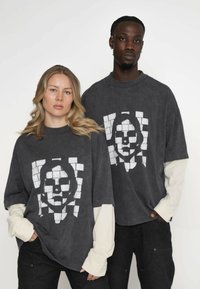 Szare, oversize'owe koszulki T-shirt wykonane z miękkiego materiału, z wyraźnym białym wzorem graficznym. Długie rękawy noszone pod spodem dodają kontrastu.