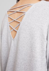 Grå tröja med V-formad rygg och korslagda band, tillverkad av ett mjukt, texturerat tyg. Designen betonar en öppen ryggstil.