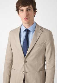 Giacca da completo beige con revers, due bottoni e tasche laterali. Indossata sopra una camicia azzurro chiaro e una cravatta blu navy. Tessuto dalla finitura liscia.