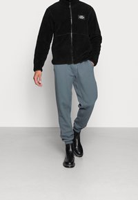 Schwarze Fleecejacke mit Reißverschluss und hohem Kragen, graue Jogginghose und schwarze Stiefeletten. Hinweis: Die Jacke hat ein Brustpatch und eine weiche Textur.