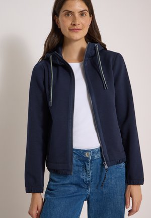 Femme portant un sweat à capuche bleu marine zippé avec cordons, chemise blanche et jean bleu, debout devant un fond clair uni.