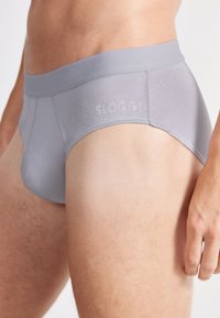 Slip Sloggi grigio realizzato in tessuto morbido con una fascia larga in vita, caratterizzato da un logo discreto nella zona della fascia. Vestibilità comoda e cuciture piatte.