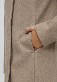 Beige vlněný kabát s texturovaným povrchem, se boční kapsou a malou kovovou štítkovou nálepkou na dolním okraji.