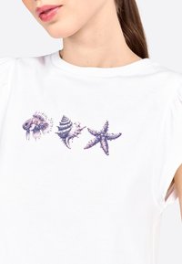 Camiseta blanca con un diseño de tres motivos marinos: una medusa, una concha y una estrella de mar, en púrpura con texturas detalladas. Mangas abullonadas.