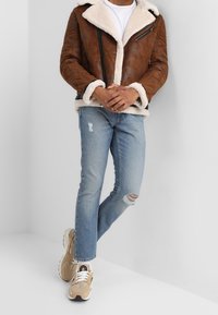 Mand iført brun shearling jakke, hvid t-shirt, slidte lyseblå jeans og beige sneakers, stående med hænderne foldet mod en hvid væg.