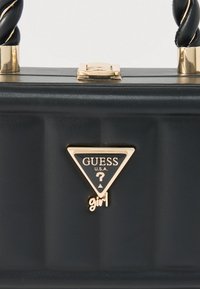 Czarna torebka z plecionymi uchwytami, złotym zapięciem oraz centralnym złotym trójkątnym logo Guess USA Girl na żebrowanej, teksturowanej powierzchni.