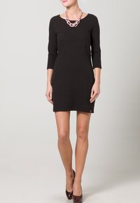 Musta istuva mekko, jossa on kolme neljäsosaa hihaa ja teksturoitu materiaali, ikoninen logo helmassa, yhdistetty paksuun vaaleanpunaiseen kaulakoruun ja tummiin korkokenkiin.