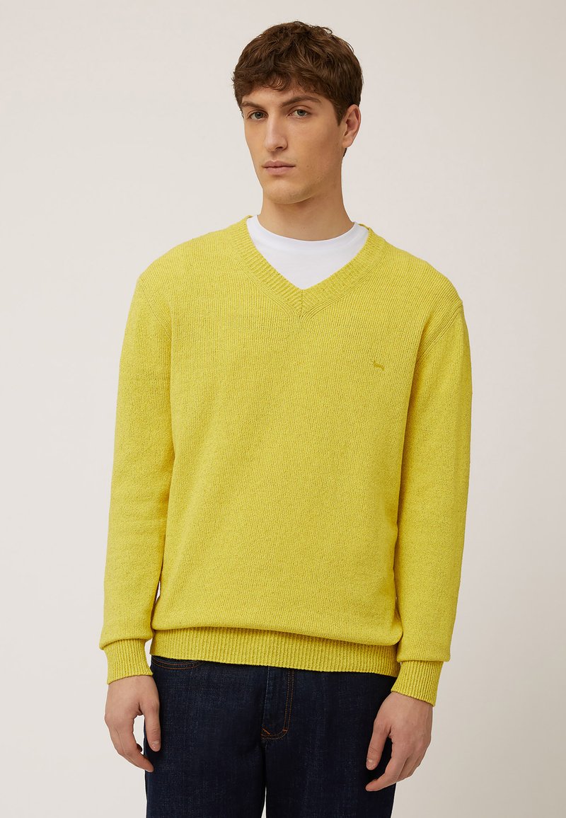 Gele V-hals trui in een gestructureerde brei met ribgebreide manchetten en zoom, gelaagd over een wit shirt, gecombineerd met donkerblauwe denimjeans.