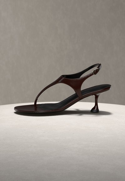 Ciemnobrązowy skórzany sandał typu slingback z cienkimi paskami, niskim obcasem i zaokrąglonym czubkiem, spoczywający na jasnej kamiennej powierzchni na szarym tle.