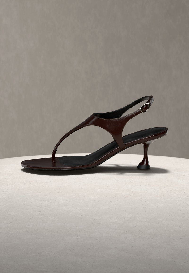 Donkerbruine leren slingback sandaal met dunne bandjes, kitten heel en afgeronde neus, rustend op een lichte stenen ondergrond tegen een grijze achtergrond.