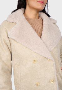Cappotto beige doppiopetto con un morbido colletto in pelliccia sintetica di colore chiaro. Presenta bottoni beige e una tasca laterale. Tessuto testurizzato.