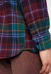 Gros plan d'une personne portant une chemise à carreaux multicolore avec des poignets boutonnés et un pantalon en velours côtelé marron, main reposant le long du corps.