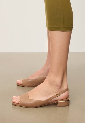 Pies con zapatos beige de tacón bajo de bloque, abiertos en la punta y con correa trasera con hebilla dorada, combinados con pantalones cortos de color verde oliva.