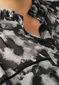 Zbliżenie czarno-szarej koszuli z wzorem, z falbaną na dekolcie i pojedynczym czarnym guzikiem blisko obojczyka.