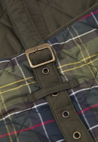 Tecido acolchoado em verde escuro, apresentando um padrão de xadrez em azul-marinho, oliva, amarelo e vermelho. Detalhe em fivela de latão e botão de pressão.