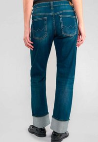 Jean en denim bleu à jambe droite, avec un ourlet retourné et des poches arrière ornées d'un motif de couture distinctif.