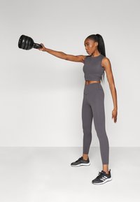 Szürke ujjatlan crop top és magas derekú leggings fekete sportcipővel párosítva. A modell egy fekete súlyzót tart, karjait kinyújtva.