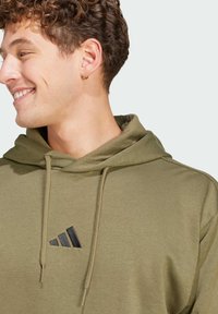 Olivovo zelená mikina vyrobená z mäkkej látky, s klokankovou kapsou a čiernym logom na hrudi. Kapucňa je stiahnutá s nastaviteľnými šnúrkami.