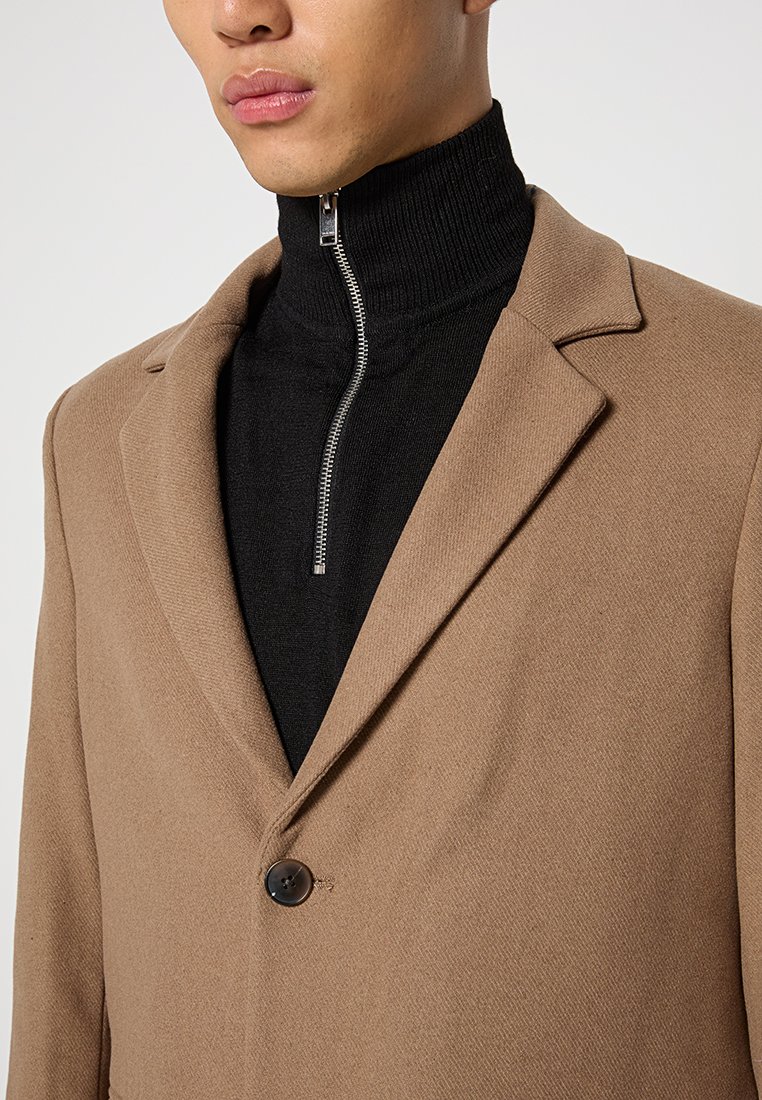 Uomo che indossa un blazer color cammello sopra un maglione turtleneck a zip nero con coste, mostrando la parte inferiore del viso e la parte superiore del torso, su uno sfondo neutro.