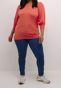 Femme portant un haut corail à manches longues, un jean skinny bleu et des baskets blanches, debout contre un fond clair uni.