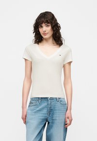 Γυναίκα με λευκό T-shirt V-λαιμόκοψης με κοντά μανίκια και ανοιχτό μπλε ψηλόμεσο τζιν, στέκεται μπροστά σε ένα μονόχρωμο φόντο.