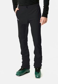 Pantalones de soft shell negros con paneles de rodilla reforzados, bolsillos con cremallera y un ajuste elástico, combinados con botas de esquí verdes para actividades invernales.