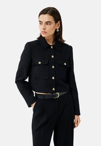 Giacca cropped nera e strutturata con maniche lunghe, due tasche frontali e bottoni dorati. Indossata con pantaloni neri a vita alta e una cintura.