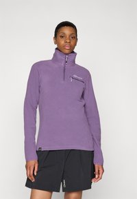 Lilaner Fleece-Pullover mit hohem Kragen, halblangem Reißverschluss und gesticktem Logo auf der Brust. Kombiniert mit schwarzen Shorts.