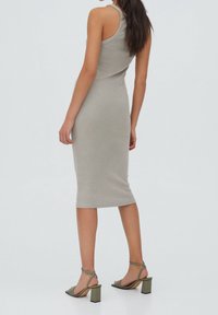 Donna in piedi che indossa un vestito beige senza maniche, lungo fino al ginocchio, e sandali con tacco a blocco verde oliva su uno sfondo semplice.