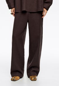 Pantalones de pierna ancha marrón oscuro, hechos de tela texturizada, combinados con zapatos de punta en un estampado de serpiente amarillo y marrón.