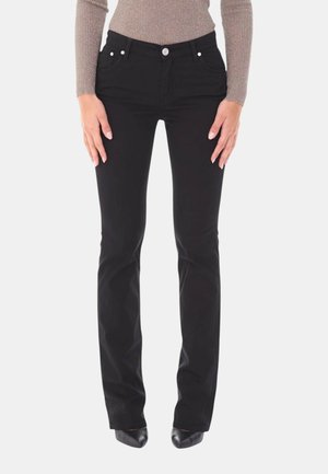 Donna che indossa pantaloni neri aderenti, maglia beige a maniche lunghe e scarpe nere con punta appuntita, in piedi con le mani lungo i fianchi.