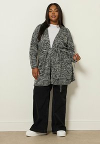 Lång grå och svart stickad cardigan med knytmidja, med djup V-ringning, långa ärmar och en lös passform. Kombineras med vida svarta byxor.