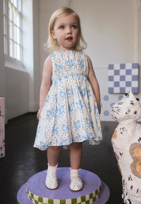 Biele šaty s modrými mašličkami a čiernymi bodkami bez rukávov s nariaseným pásom. Biele sandále s dekoratívnymi remienkami. Stojí na okrúhlom podstavci.