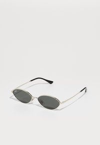 Gafas de sol ovaladas con lentes grises, montura de metal dorado delgada, puntas de patilla negras y un diseño minimalista. Presenta el logotipo en la lente.